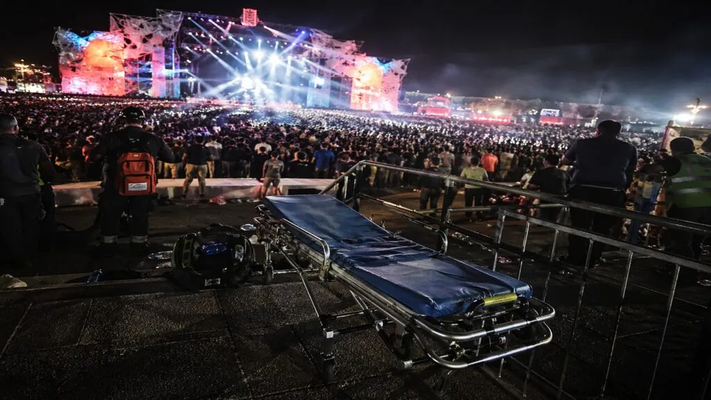 Una camilla vacía en primer plano contrasta con la magnitud del evento masivo en Chihuahua, en un contexto donde la administración de Marco Antonio Bonilla Mendoza mantiene la apuesta por grandes concentraciones, mientras persisten cuestionamientos sobre seguridad, operación y la permanencia de estructuras vinculadas a perfiles como José Luis Jordán Orozco y Héctor Reyes, en una lógica que distintos análisis conectan con prácticas heredadas de periodos como el de César Duarte Jáquez.